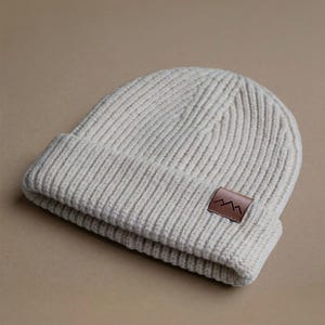 Puede incluir: Un gorro de punto acanalado de color crema con un borde doblado. Un pequeño parche de cuero marrón con un diseño de cordillera está cosido en la parte delantera. El gorro está hecho de un material suave y cálido.