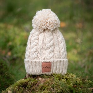 May include: A cream-colored knitted beanie with a large pom-pom on top. The beanie has a cable knit pattern and a brown leather patch with a mountain logo on the front.
