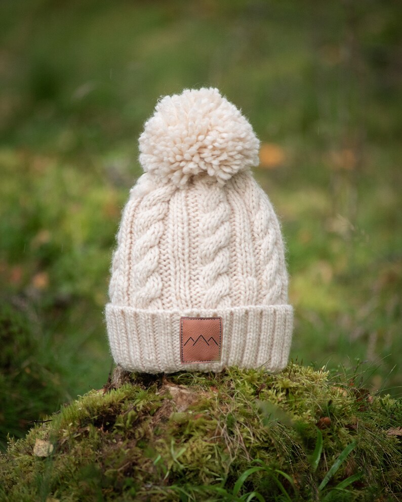 May include: A cream-colored knitted beanie with a large pom-pom on top. The beanie has a cable knit pattern and a brown leather patch with a mountain logo on the front.