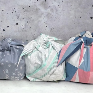 Puede incluir: Tres paquetes de envoltura de regalo de tela con diferentes patrones. Los paquetes están atados con un nudo en la parte superior. Los paquetes están sobre un fondo gris.