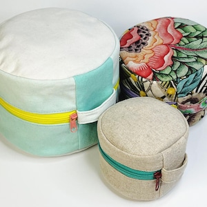 Op de afbeelding: Drie ronde opbergcontainers van stof met ritsen. De grootste container is lichtblauw en wit met een gele rits. De middelste container is bedekt met een bloemenprint met groene bladeren en roze bloemen. De kleinste container is beige met een turquoise rits.