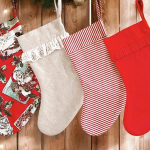 Puede incluir: Cuatro calcetines de Navidad colgados en una pared de madera. Los calcetines están hechos de diferentes telas y tienen varios diseños. Un calcetín es rojo con una ilustración navideña vintage, otro es de lino beige con un borde de encaje, un tercero es de rayas rojas y blancas, y el último es rojo sólido con un puño de punto.