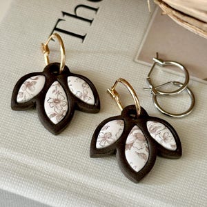 Peut inclure: Une paire de boucles d'oreilles avec des montures en forme de feuille brun foncé et des incrustations blanches à motifs floraux suspendues à des anneaux dorés, accompagnées d'une paire d'anneaux argentés sur une surface texturée.