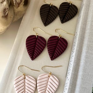 Peut inclure: Trois paires de boucles d'oreilles pendantes en forme de feuille avec des crochets dorés. La paire supérieure est marron foncé, la paire centrale est bordeaux profond et la paire inférieure est rose poudré. Chaque boucle présente un motif nervuré.
