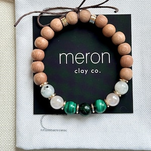 May include: A bracelet with wooden beads, white and green gemstones, and gold-coloured spacers. The bracelet is presented on a black card with the text "meron clay co." and tied with a brown string.