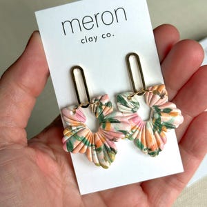 以下が含まれることがあります： ゴールドカラーの金具が付いた、花柄のイヤリング。イヤリングはピンク、緑、オレンジの色合いの花の形をしています。「meron clay co.」の文字が入った白いカードに付けられています。