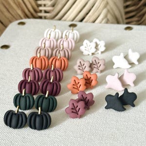 May include: A collection of stud earrings featuring miniature pumpkins in various colours, leaf shapes in autumn tones, and ghost figures in white, pink, and black. Each piece has a small gold-toned stem or detail, arranged in rows on a textured surface.