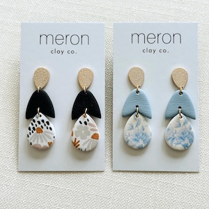 May include: Two pairs of handmade polymer clay earrings. One pair features black, white, and floral patterns, while the other has blue and white floral designs. Each earring has a gold-coloured top and is displayed on a white card with the text "meron clay co."