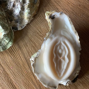 May include: A white candle shaped like a vulva, nestled inside an oyster shell. The candle has detailed features and sits on a textured wooden surface. Two other oyster shells are visible in the background.