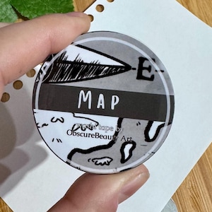 Peut inclure: Un autocollant de carte noir et blanc avec le mot "MAP" imprimé en blanc sur une bande noire. L'autocollant est rond et a une bordure blanche. Le texte "Washi tape by ObscureBeauty Art" est imprimé en blanc sur fond noir en bas de l'autocollant.