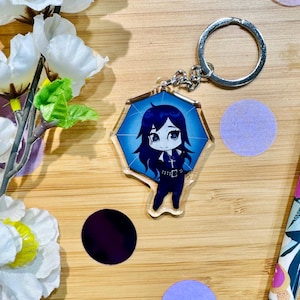 May include: A blue and white anime character keyring with a silver key ring. The character is a girl with long black hair and a black outfit. The keyring is shaped like a diamond.