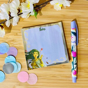 May include: A notepad with the word "Notes" and an illustration of a woman with green hair. A floral pen and colorful confetti are next to the notepad. White flowers are in the background. The notepad and pen are on a wooden surface.