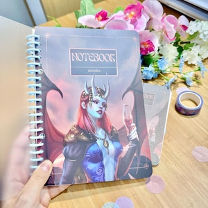 Könnte beinhalten: Ein spiralgebundenes Notizbuch mit einem bunten Cover, das eine Frau mit roten Haaren und Hörnern zeigt, die ein blaues und violettes Kleid trägt und ein Glas Wein hält. Das Cover trägt auch den Text "NOTEBOOK" und "MIZORA".