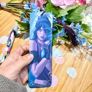 Könnte beinhalten: Ein Lesezeichen mit einer Frau mit blauen Haaren und einem violetten Outfit, die ein blaues Juwel hält. Das Lesezeichen hat einen goldenen Rand und einen blauen Hintergrund.