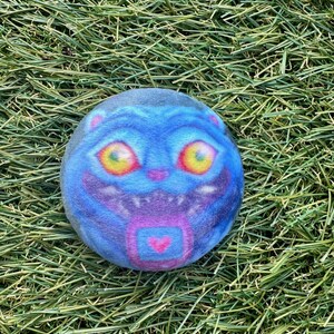 May include: A round, blue plush toy with a cartoon cat face. The cat has large yellow eyes, a purple mouth, and a pink heart design. The toy is set against a background of green grass.