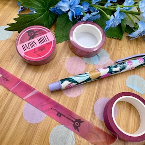 May include: An assortment of stationery items, including washi tape rolls and a pen, are arranged on a wooden surface. The washi tape rolls are a deep pink color, one with the text "HAZBIN HOTEL". The pen has a floral design. Blue flowers and confetti are in the background.