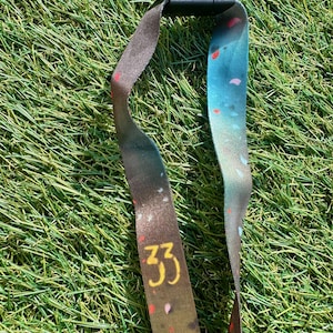 Könnte beinhalten: Ein Schlüsselband mit schwarzem Verschluss und gemustertem Band. Das Band zeigt einen Farbverlauf aus Braun, Blau und Weiß mit der goldenen Zahl "33". Kleine, farbige Formen sind über das Band verstreut.