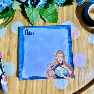 May include: Blue and white patterned notepad with the word "Notes" written in black ink. The notepad features a drawing of a woman with long blonde hair, wearing a blue and green dress. The woman has pointed ears and is holding a glowing orb.