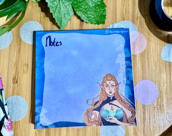 May include: Blue and white patterned notepad with the word "Notes" written in black ink. The notepad features a drawing of a woman with long blonde hair, wearing a blue and green dress. The woman has pointed ears and is holding a glowing orb.