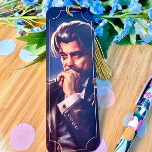 Puede incluir: Un marcapáginas azul marino con un retrato de un hombre con bigote y expresión pensativa. El marcapáginas tiene una borla dorada y está rodeado de flores azules y confeti. También se ve un bolígrafo con un diseño floral.