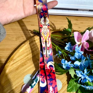 May include: A colourful patterned lanyard with a silver metal clasp. The lanyard features a repeating pattern of bats, moons, and floral designs in shades of red, blue, gold, and white.