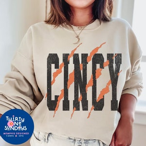 May include: A beige sweatshirt with the word "CINCY" printed in black with orange claw marks behind the letters.