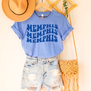 May include: A light blue t-shirt with the word "Memphis" repeated three times in a retro, wavy font. The shirt is hanging on a wooden rack with a straw hat and a woven bag.