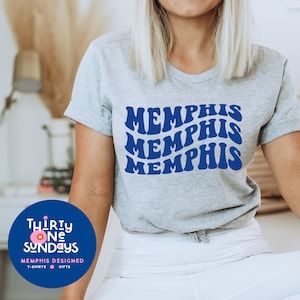 May include: A gray t-shirt with the word "Memphis" repeated three times in a blue retro font. The shirt is being worn by a person. The image also includes a logo for "Thirty One Sundays" with a pink circle and the text "Memphis Designed, T-shirts, Gifts."