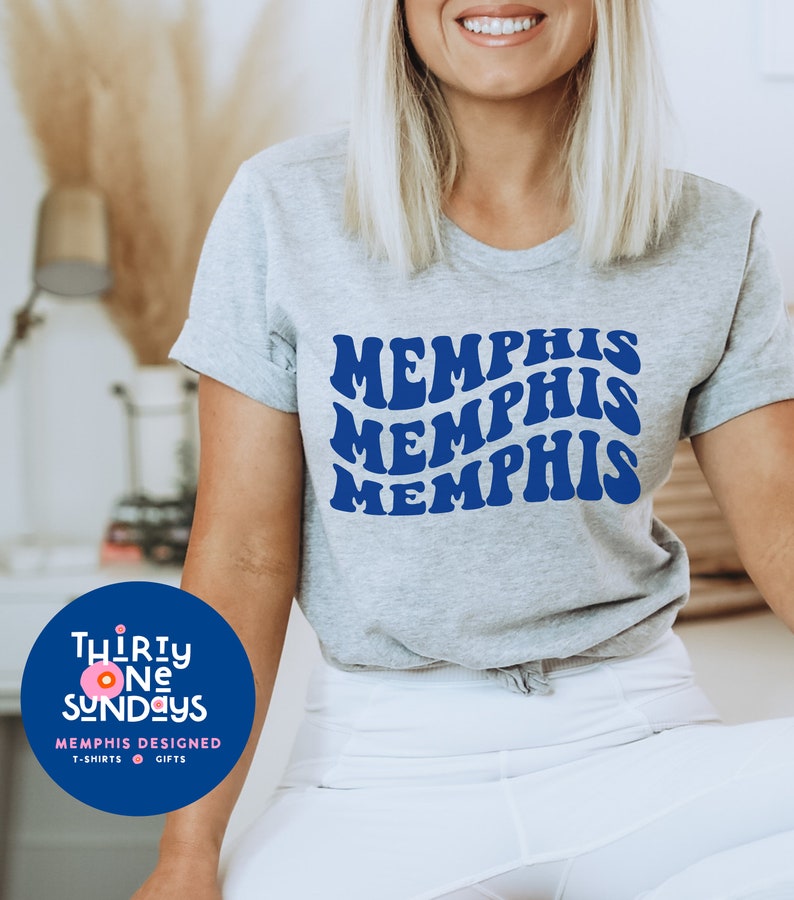 May include: A gray t-shirt with the word "Memphis" repeated three times in a blue retro font. The shirt is being worn by a person. The image also includes a logo for "Thirty One Sundays" with a pink circle and the text "Memphis Designed, T-shirts, Gifts."