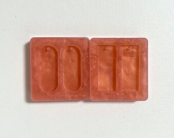 Puede incluir: Dos moldes de silicona rosa para hacer pendientes de resina. Un molde tiene dos formas ovaladas con un agujero en la parte superior, el otro molde tiene dos formas rectangulares.