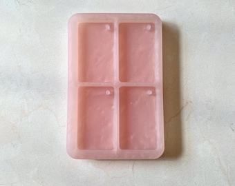 Puede incluir: Un molde rectangular de silicona rosa claro con cuatro cavidades rectangulares. Cada cavidad tiene un pequeño agujero en la parte superior para colgar. El molde está diseñado para crear objetos rectangulares pequeños.