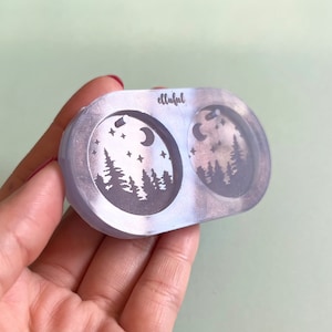 May include: A clear silicone mold with two circular cavities. Each cavity features a design of a crescent moon, stars, and a silhouette of a forest. The mold is labeled "ellahful".