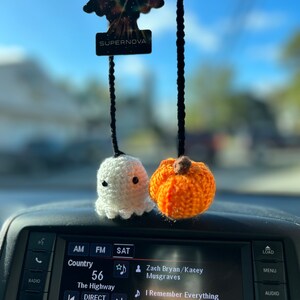 May include: Crocheted Halloween car charms. A white ghost and an orange pumpkin hang from black strings. The car radio displays the text "Country 56" and the song "I Remember Everything". A "SUPERNOVA" air freshener hangs above.