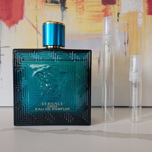 May include: A blue glass bottle of Versace Eros Eau de Parfum with a gold cap. The bottle is sitting on a white surface next to two smaller clear glass bottles with silver caps.