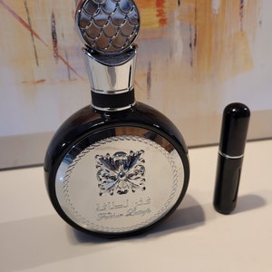 May include: A dark blue perfume bottle with a silver cap and a matching travel-size spray bottle. The perfume bottle features a silver emblem with Arabic script and a decorative silver cap. The background is an abstract painting.