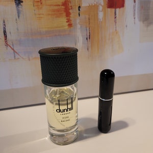 May include: A clear glass bottle of Dunhill London Icon Racing cologne with a black cap and a black and silver atomiser. The bottle is sitting on a white surface next to a smaller black travel-sized atomiser.