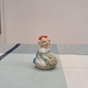 May include: A small ceramic figurine of a seated elf with red hair, wearing a blue and white outfit. The elf is holding a basket of green objects. The figurine is on a checkered blue and white surface.