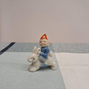 May include: A small ceramic figurine of a gnome riding a white rabbit. The gnome is dressed in blue with an orange hat. The rabbit is white with detailed features. The figurine is set against a light blue and white checkered background.