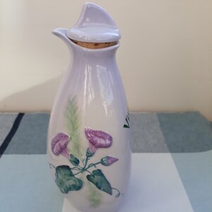 May include: A lavender ceramic bottle with a cork stopper and a floral design. The bottle has a spout and is decorated with a vine of green leaves and purple morning glory flowers.