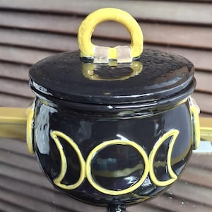 May include: A black ceramic cauldron with a matching lid featuring a yellow handle. The exterior of the pot is decorated with a yellow triple moon symbol, consisting of a central full circle flanked by two crescent moons, and includes three small feet.
