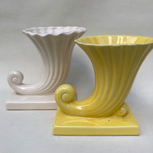 May include: Two ceramic vases in the shape of a horn. One vase is white and the other is yellow. Both vases have a fluted design and a curled handle.