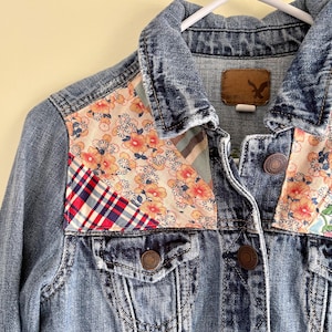 May include: A denim jacket with patchwork floral and plaid fabric accents. The jacket features a classic collar, button-down front, and two chest pockets. The patchwork includes red, blue, and cream floral patterns.