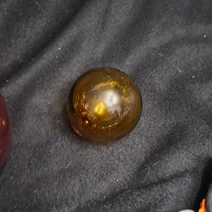 May include: A spherical glass float with a translucent amber-brown color. The surface of the orb is smooth and reflective, showing a bright highlight from a light source. The object is circular in shape and appears to be made of blown glass.