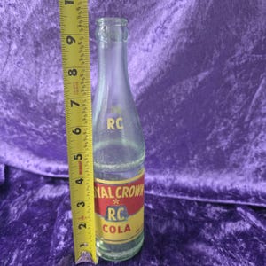 May include: A clear glass Royal Crown RC Cola bottle with a red and yellow label featuring the text 'ROYAL CROWN RC COLA'. A yellow measuring tape is positioned vertically next to the bottle, indicating a height of approximately 9.5 inches.