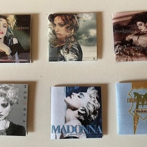 May include: Six square prints featuring various Madonna album covers arranged in two rows. The images display portraits of the artist and the text Madonna, along with the Immaculate collection album cover featuring a gold crest on a light blue background.