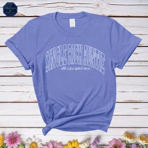 Puede incluir: Una camiseta azul claro con el texto "Single Rich Auntie with a few spoiled nieces" en letras blancas delineadas.