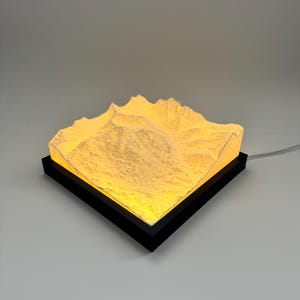 Puede incluir: Una escultura cuadrada e iluminada de una cordillera. La cordillera es de color blanquecino texturizado y emite un brillo amarillo cálido. Se encuentra dentro de un marco cuadrado negro. Un cable de alimentación se extiende desde el marco.