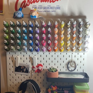 Könnte beinhalten: Eine weiße Lochwand mit farbigen Garnrollen, einem Foto und kleinen Accessoires. Das Garn ist in einem Farbverlauf angeordnet. Im Hintergrund ist ein Vintage-Poster des Films "Casablanca" zu sehen.