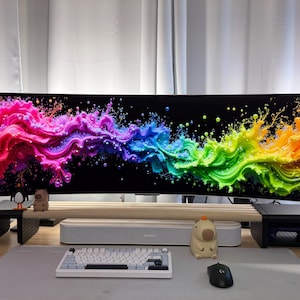 Puede incluir: Un monitor de ordenador ultra ancho curvo que muestra un diseño abstracto vibrante y colorido. La configuración incluye un teclado blanco, un ratón y figuras decorativas. El monitor está soportado por soportes negros y una estantería de madera.