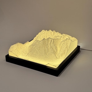 Puede incluir: Una cordillera impresa en 3D, iluminada desde dentro, sobre una base rectangular negra. El modelo de montaña es de color beige claro, con crestas y valles detallados. La base tiene un diseño elegante y moderno.